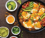 Huevos Rancheros in a skillet with avocado, and various toppings on a by wooden table