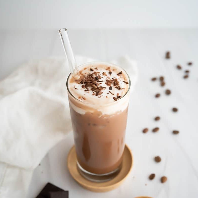 A tall glass of iced coffee or milk coffee topped with whipped cream and chocolate shavings, with a clear straw, set on a small wooden coaster on a light surface with scattered coffee beans and a white cloth nearby.
