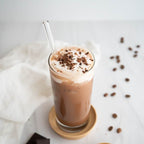 A tall glass of iced coffee or milk coffee topped with whipped cream and chocolate shavings, with a clear straw, set on a small wooden coaster on a light surface with scattered coffee beans and a white cloth nearby.