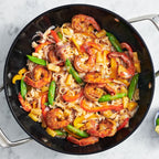 A top-down view of a black cast iron skillet filled with Jerk shrimp and rice noodles, with snow peas, red and yellow peppers, and served with small bowls of chopped parsley and avocado on a white marble surface.