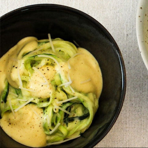 A bowl of Keto Modern Mac with zucchini ribbons and a sprinkle of black pepper, photographed from above on a light textured fabric background.