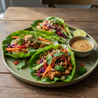 Three fresh lettuce wraps filled with seasoned minced meat, carrots, purple cabbage, peanuts, and herbs, served on a round green plate with a small bowl of dipping sauce on a rustic wooden table.