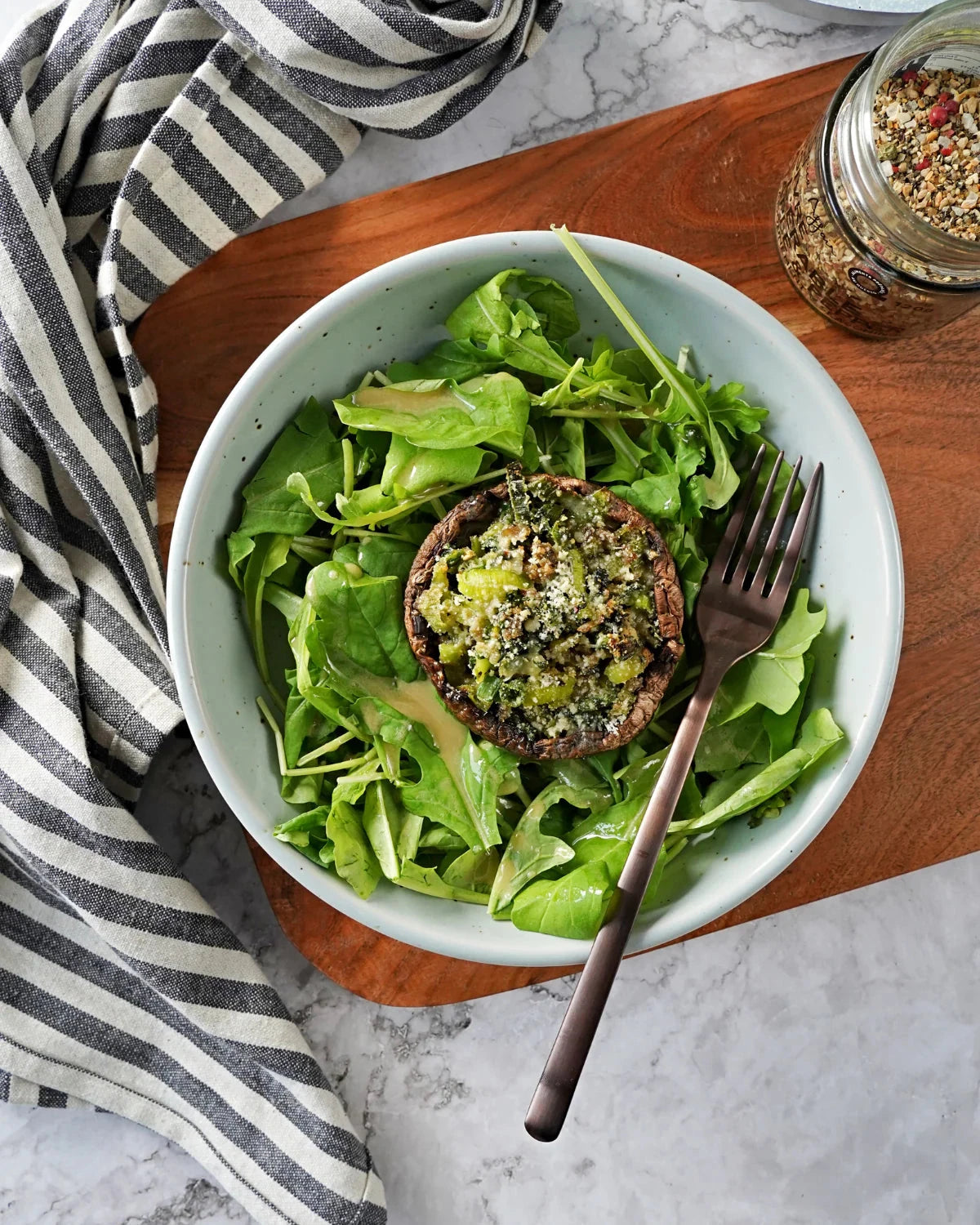 Maple & Pepper Seasoning sprinkled over a stuffed mushroom on fresh green salad, with the seasoning jar and fork arranged on a wooden board