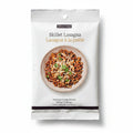 A package of Epicure Skillet Lasagna Seasoning centered on a light marble surface, surrounded by pasta, garlic, herbs, and a small bowl of dried herbs.