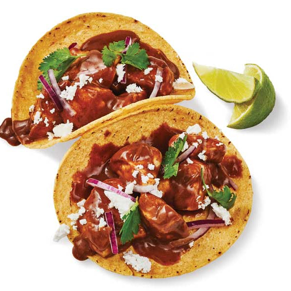 Two soft corn tacos filled with cooked meat in Epicure Mole in Minutes sauce, topped with crumbled white cheese, red onion, and cilantro, served on a white background with lime wedges beside them.