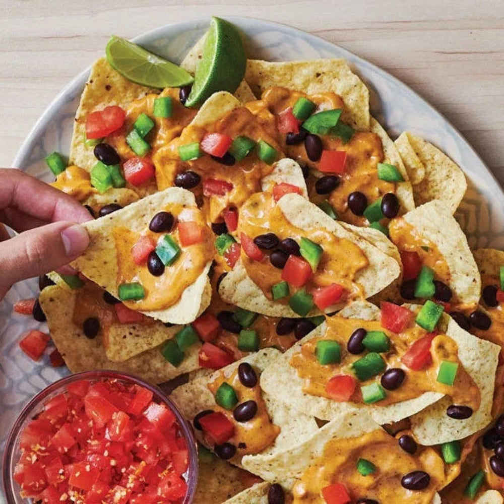Ooey Gooey Queso Hot Dip Mix with loaded nachos topped with melted cheese, black beans, tomatoes, peppers, lime wedges, and salsa on the side