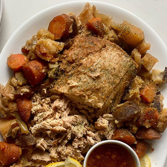 A plated meal featuring a Portuguese Chicken Seasoning on pork roast  with carrots, potatoes, and shredded stuffing, served on a white oval plate with a small dipping sauce cup and stacked plates with forks on a light tabletop.