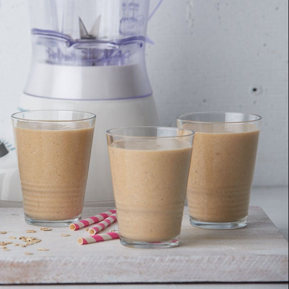 Three glasses of Pumpkin Pie Spice oatmeal milkshake with rolled oats in a glass jar, a white and purple blender in the background, and two pink striped straws on a white surface.