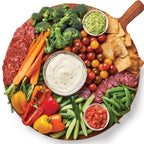 A large round wooden serving board filled with a charcuterie-style spread, photographed from above on a white background. Visible items include sliced salami, broccoli, carrots, bell peppers, cucumber, green beans, tortilla chips, a bowl of Epicure Queso Blanco Hot Dip, a bowl of salsa, and a small bowl of green dip.