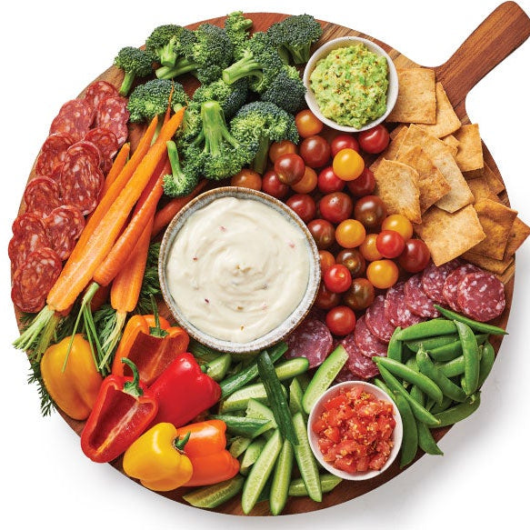 A large round wooden serving board filled with a charcuterie-style spread, photographed from above on a white background. Visible items include sliced salami, broccoli, carrots, bell peppers, cucumber, green beans, tortilla chips, a bowl of Epicure Queso Blanco Hot Dip, a bowl of salsa, and a small bowl of green dip.