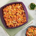 Queso Blanco Rose Pasta baked in a silicone purple baking dish, with a smaller bowl of the same pasta on a light marble surface. A green napkin, parsley garnish, a glass of water, and forks are arranged around the food.