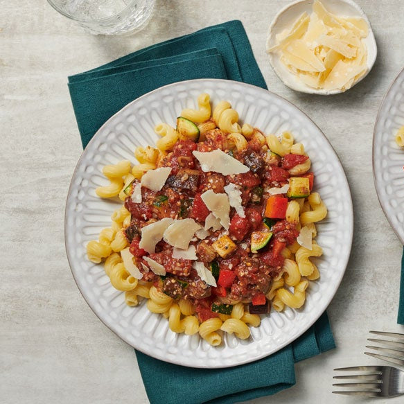 A white plates of Ratatouille Pasta with shavings of cheese, arranged on a light tabletop with teal napkins, forks, and a small bowl of cheese.