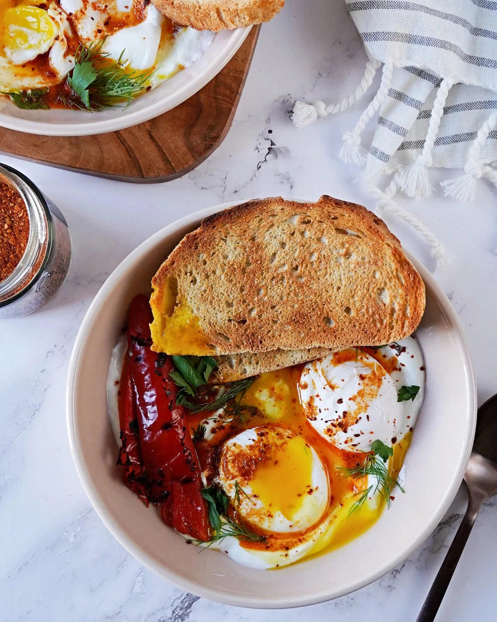 Roasted Garlic & Sun Dried Tomato Seasoning beside a bowl of eggs with roasted red peppers dill and toasted bread on a marble table