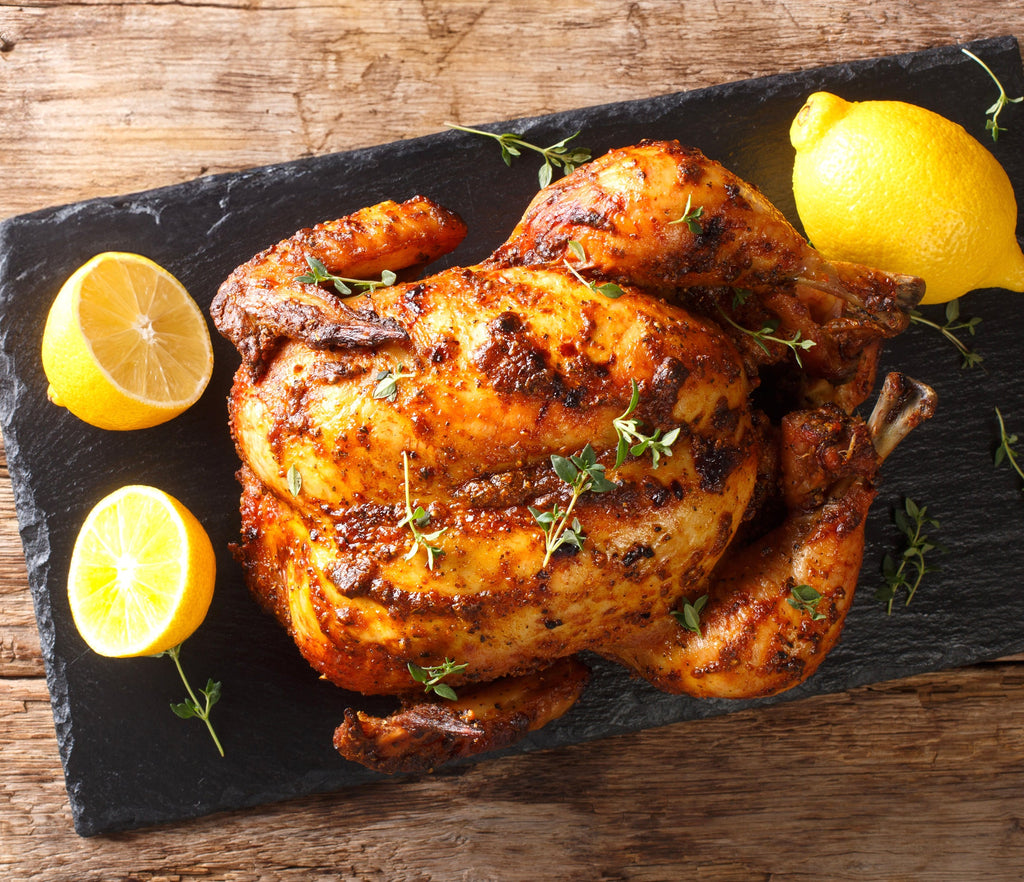 Oven roasted Epicure Rotisserie Chicken with crispy golden-brown skin sits on a dark rectangular serving board, garnished with fresh thyme, with lemon halves placed on both sides, on a rustic wooden table.