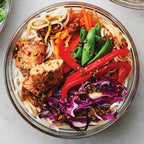 A clear glass bowl filled with colorful noodles, seasoned chicken pieces, red bell peppers, green snap peas, purple cabbage, and carrots, dressed wtih Epicure Sesame Ginger Dressing, photographed from above on a light marble surface. 