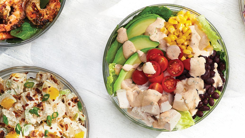 Salad bowl dressed with Epicure Southwest Ranch alongside potato salad and barbecued shrimp on a white background