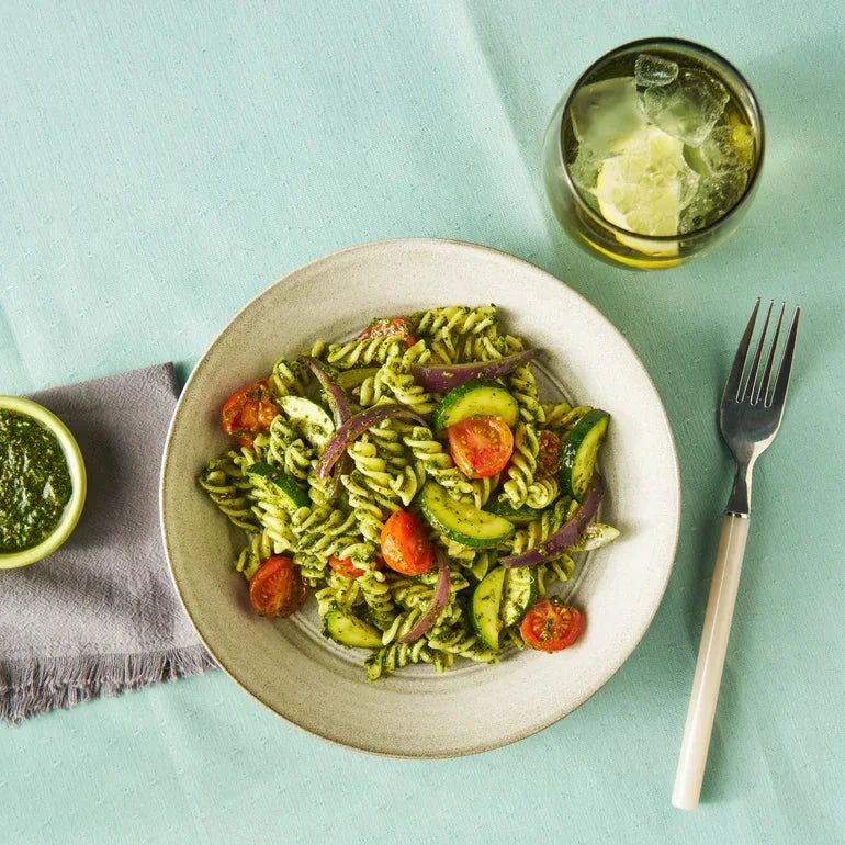 Super Green Pesto pasta dish with vegetables on a white plate, accompanied by a glass of white wine and a small bowl of pesto on a light blue background.