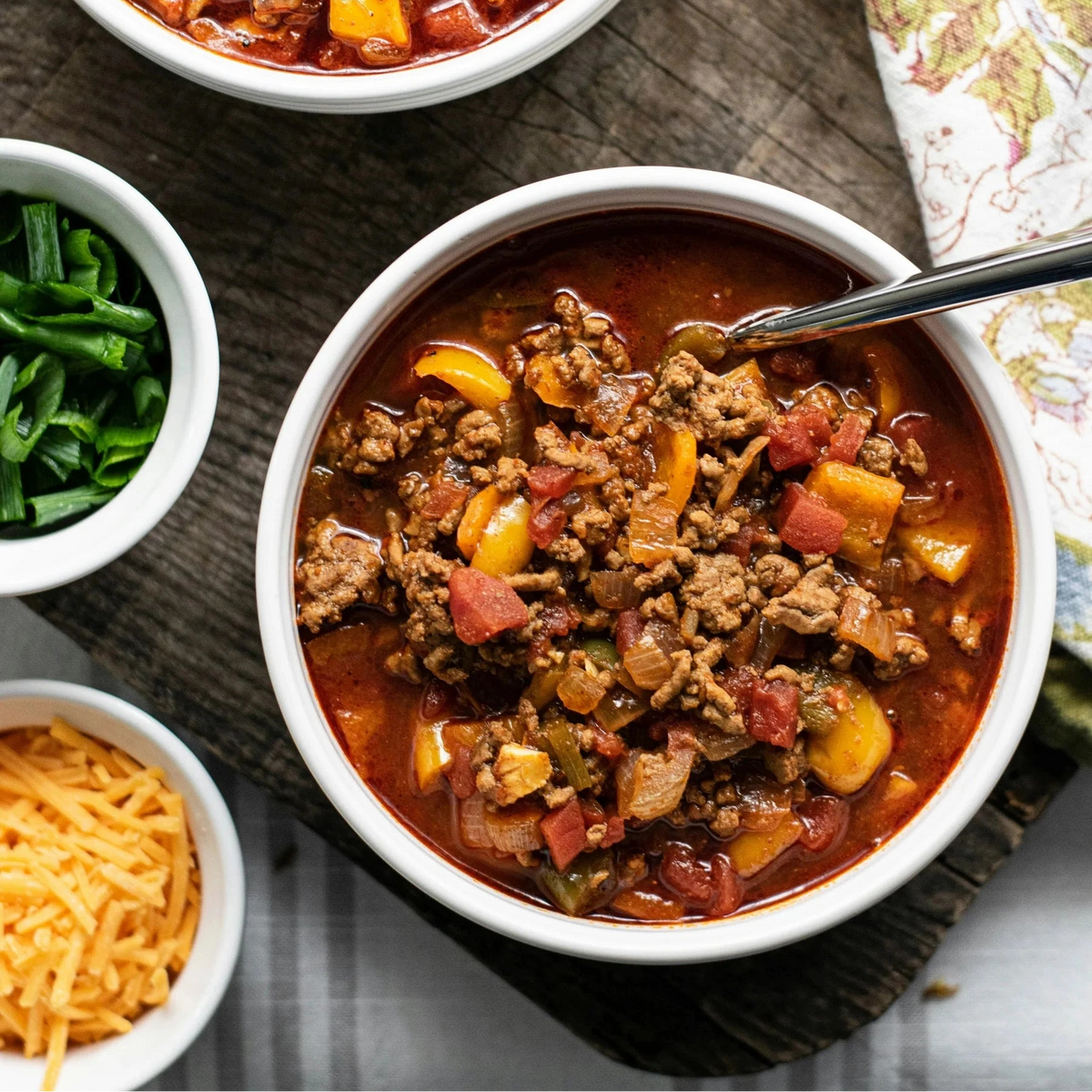 Two bowls of chili with spoons on a wooden board, accompanied by small bowls of greens and cheese.