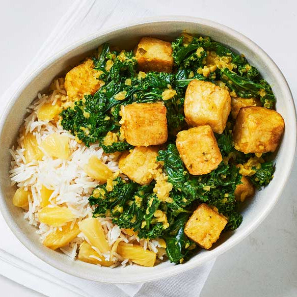 A top-down view of a white bowl filled with fried tofu cubes, green broccoli, and white rice, with chopsticks resting on the right side on a light marble surface.