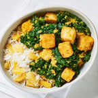 A top-down view of a white bowl filled with fried tofu cubes, green broccoli, and white rice, with chopsticks resting on the right side on a light marble surface.