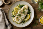 A cooked fish fillet served on a ceramic plate with lemon slices, greens, and dill, set on a rustic wooden table with small bowls and a napkin nearby.
