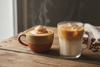 A ceramic mug with a brown and beige glazed finish holds a steaming latter topped with a dollop of whipped cream, set beside a clear glass mug filled with an iced latte on a rustic wooden table, with scattered coffee beans in the background.
